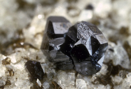 Ein Mineral der Davidit-Crichtonit-Gruppe| Aufgrund der Kristallform, Verzwillingung, Korrosionsspuren, starker Streifung und der Paragenese mit Magnetit und Hämatit dürfte es sich bei diesen Kristallen um ein Mineral der Davidit-Crichtonit-Gruppe handeln, möglicherweise einen Crichtonit. (Aggregat 3,5mm). Ähnliche schwarze Kristalle treten vereinzelt in der Lercheltinizone auf, die jedoch gegen Ilmenit makroskopisch z.T. wohl nicht sicher abzugrenzen sind. Frischer Ilmenit scheint jedoch nicht gemeinsam mit Magnetit oder Hämatit vorzukommen 