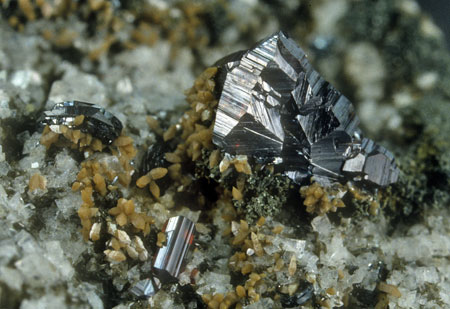 Rutile unterschiedlichen Bildungsalters| Auf einem Aggregat frühgebildeter kurzprismatischen Rutilkristalle (8 mm Durchmesser) sind nacheinander Eisenrosen, Anatas und Chlorit aufgewachsen. An der unteren Bildkante sind jüngere prismatische Rutile sichtbar, die z.T. auf Adular aufgewachsen sind 